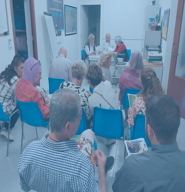 Reunión en la sede de la Asociación DESAL con varias personas sentadas en sillas azules, participando en un encuentro informativo o asamblea. Al fondo, tres personas coordinan la actividad desde una mesa con ordenadores y documentación.
