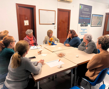 Un grupo de mujeres está reunido alrededor de una mesa en una sala, participando en una reunión o taller sobre adicciones. Hay documentos, comida ligera y un cartel informativo al fondo.