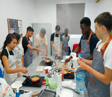 Grupo de jóvenes participando en una clase de cocina, todos con delantales y cocinando en varias sartenes sobre mesas con placas eléctricas. El ambiente es colaborativo y formativo.
