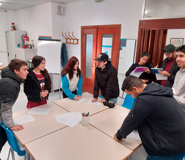Grupo de jóvenes reunidos alrededor de una mesa en una sala, participando en una actividad con hojas de papel. Algunos escriben mientras otros conversan o observan. Ambiente informal y colaborativo.